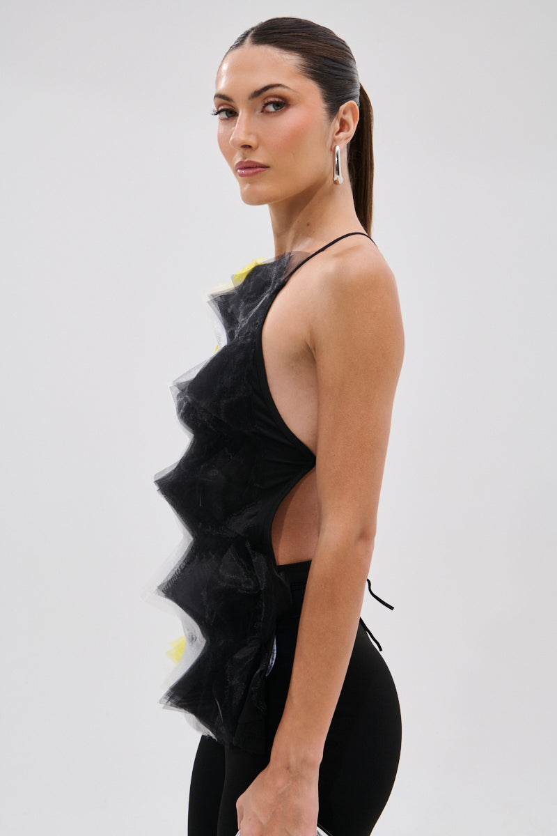 A woman with long dark hair in a sleek ponytail poses sideways against a plain light background, wearing the PIPPA MESH BLOUSE featuring dramatic ruffled detailing and black pants.