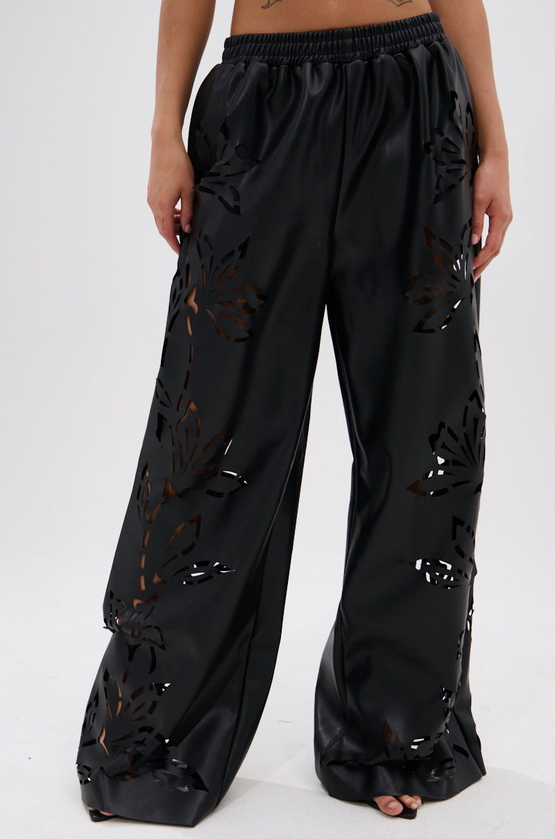 A person stands barefoot on a white background, wearing the GOING CRAZY PU CUTOUT PALAZZO PANT—black, faux leather pants with floral stencil cut-outs, elastic waistband, and wide legs.