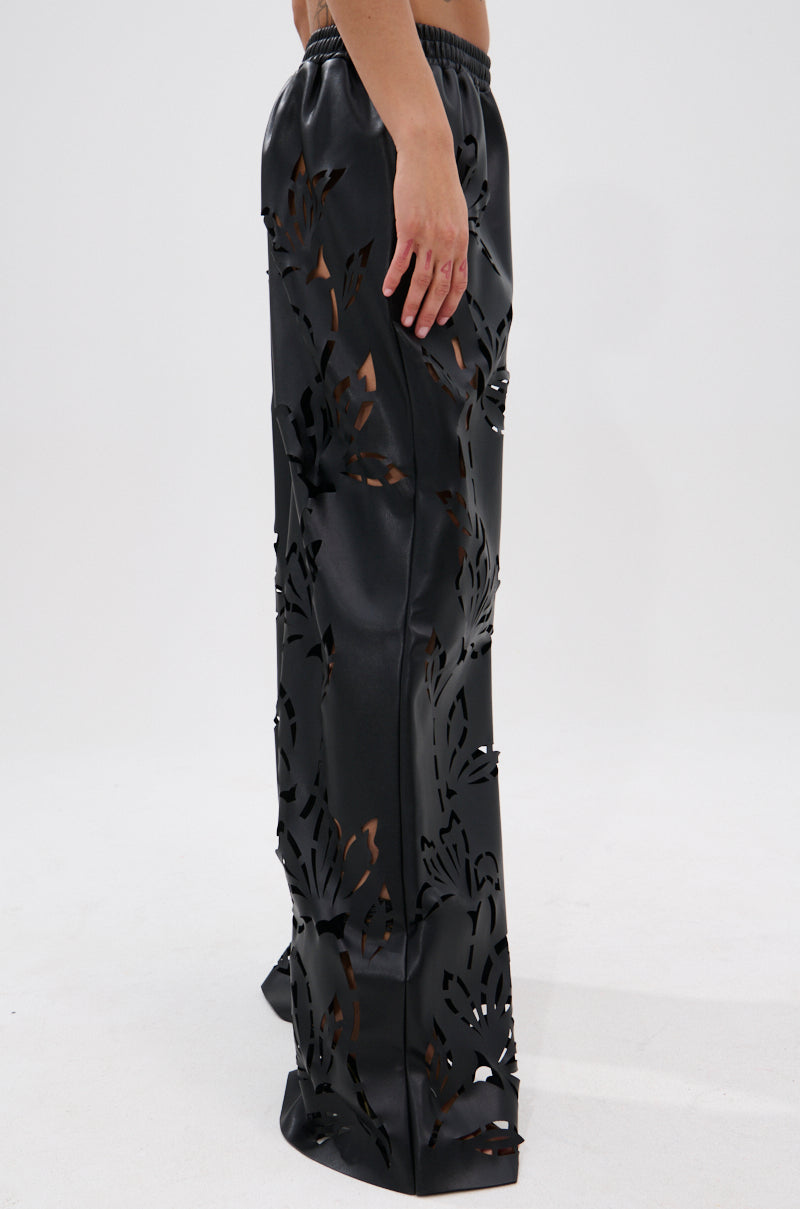 A person models the GOING CRAZY PU CUTOUT PALAZZO PANT, featuring loose-fitting, black PU material with detailed floral stencil cut-outs, an elastic waistband, and a touch of shine against a plain white backdrop.