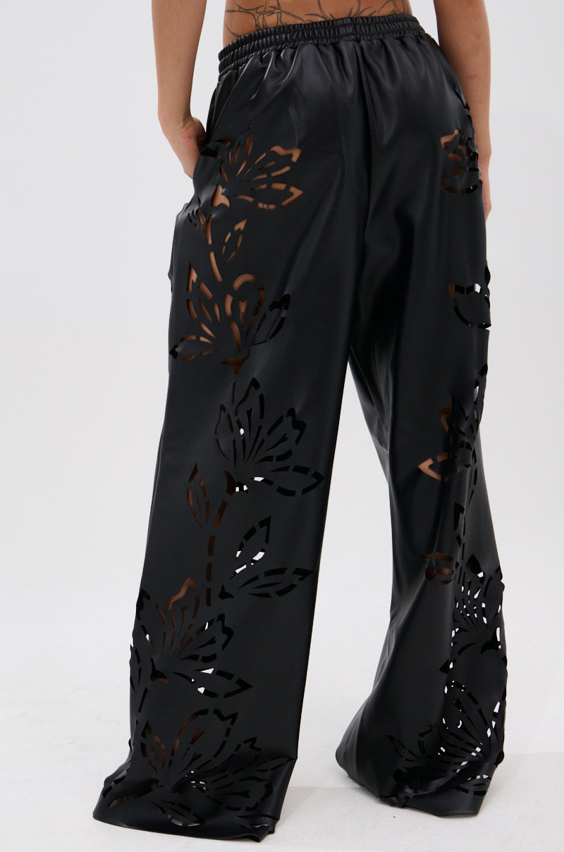A person wears the GOING CRAZY PU CUTOUT PALAZZO PANT—loose black faux leather pants featuring floral stencil laser-cut designs that reveal skin. The image, cropped from the waist down, highlights the pants’ unique back and side details.