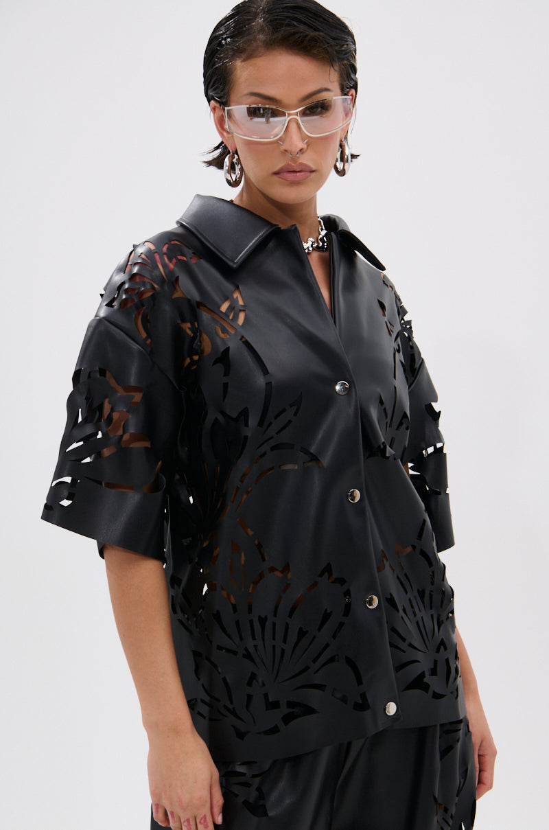 A person wears clear glasses, chunky earrings, and the GOING CRAZY PU CUTOUT BUTTON DOWN TOP—a black, floral, short-sleeve oversized shirt—paired with matching pants against a plain light background.