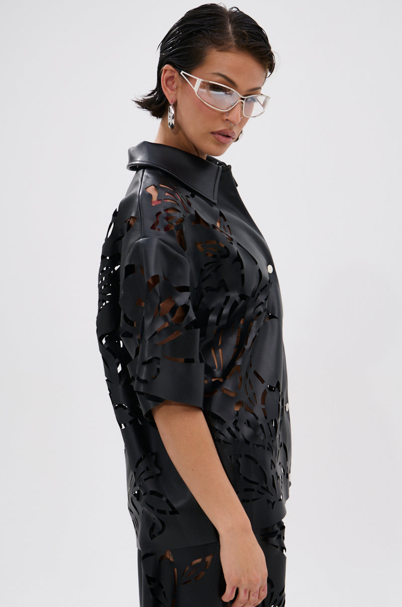 A woman with short dark hair and clear glasses wears the GOING CRAZY PU CUTOUT BUTTON DOWN TOP, a black faux leather short-sleeved shirt with floral laser-cut patterns. She stands against a plain white background, gazing downward to the side.