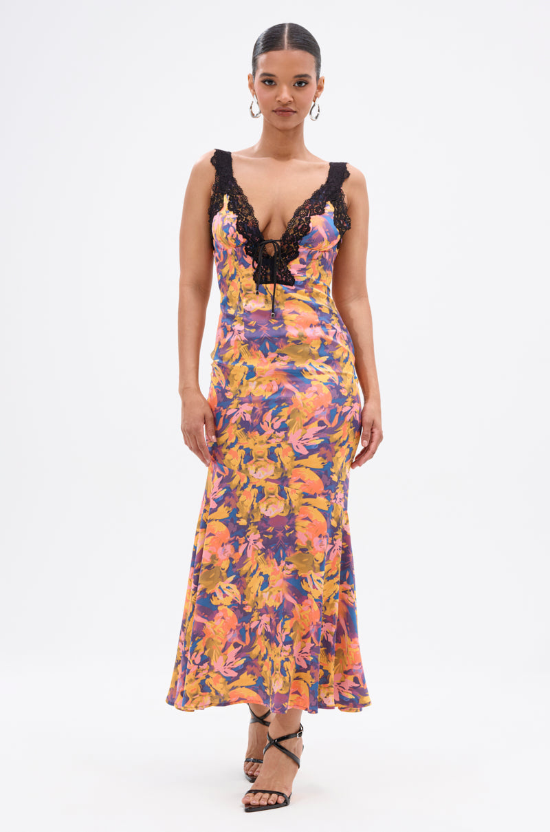A woman stands against a white background wearing the RYAN FLORAL PRINTED SATIN MIDI DRESS, featuring a sleeveless design with floral print, plunging neckline, black lace trim, and thin straps. She has slicked-back hair, hoop earrings, and strappy heels.
