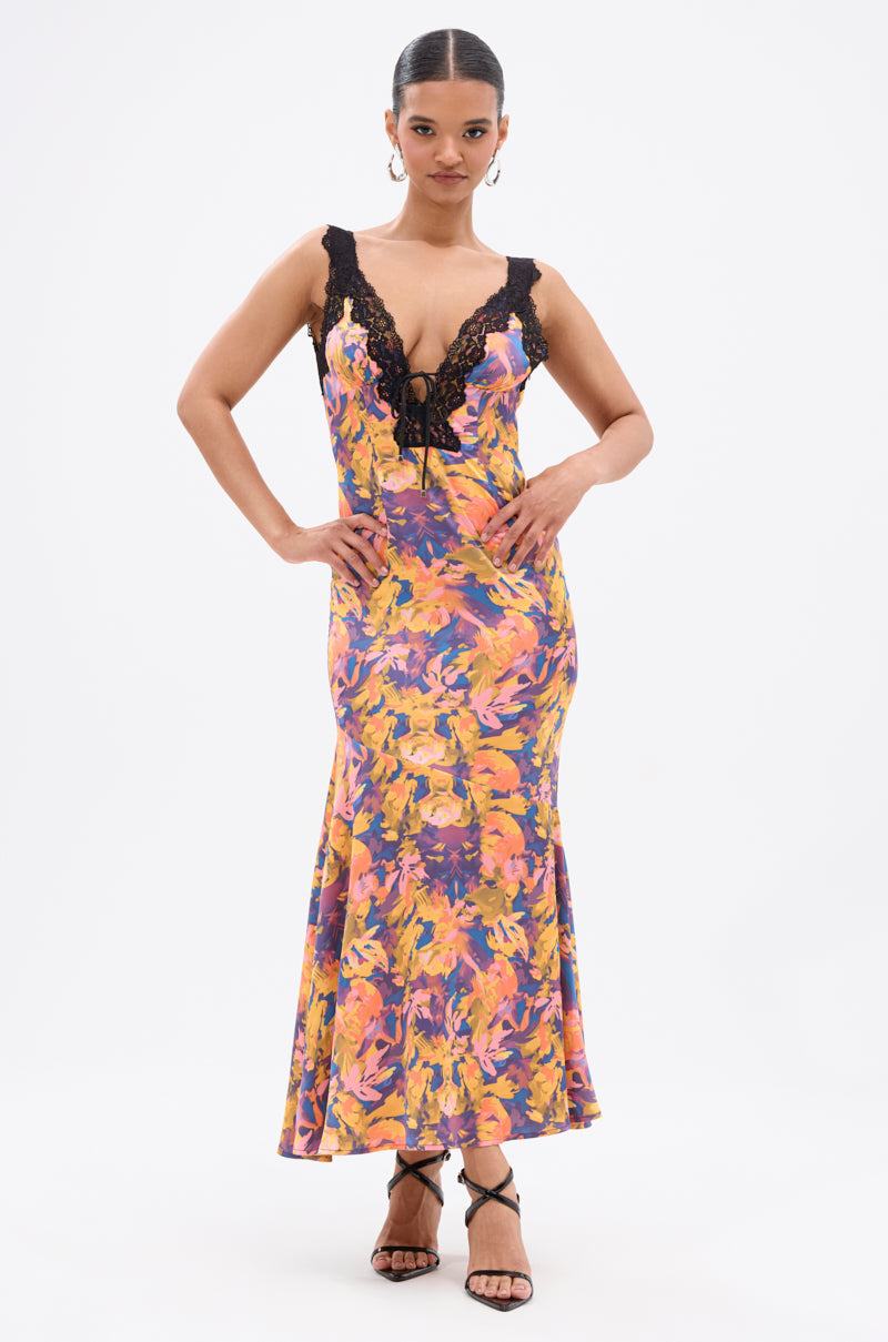 A woman poses in the RYAN FLORAL PRINTED SATIN MIDI DRESS, featuring a sleeveless floral print design with black lace details and a plunging neckline. She pairs it with strappy black heels against a plain white background.