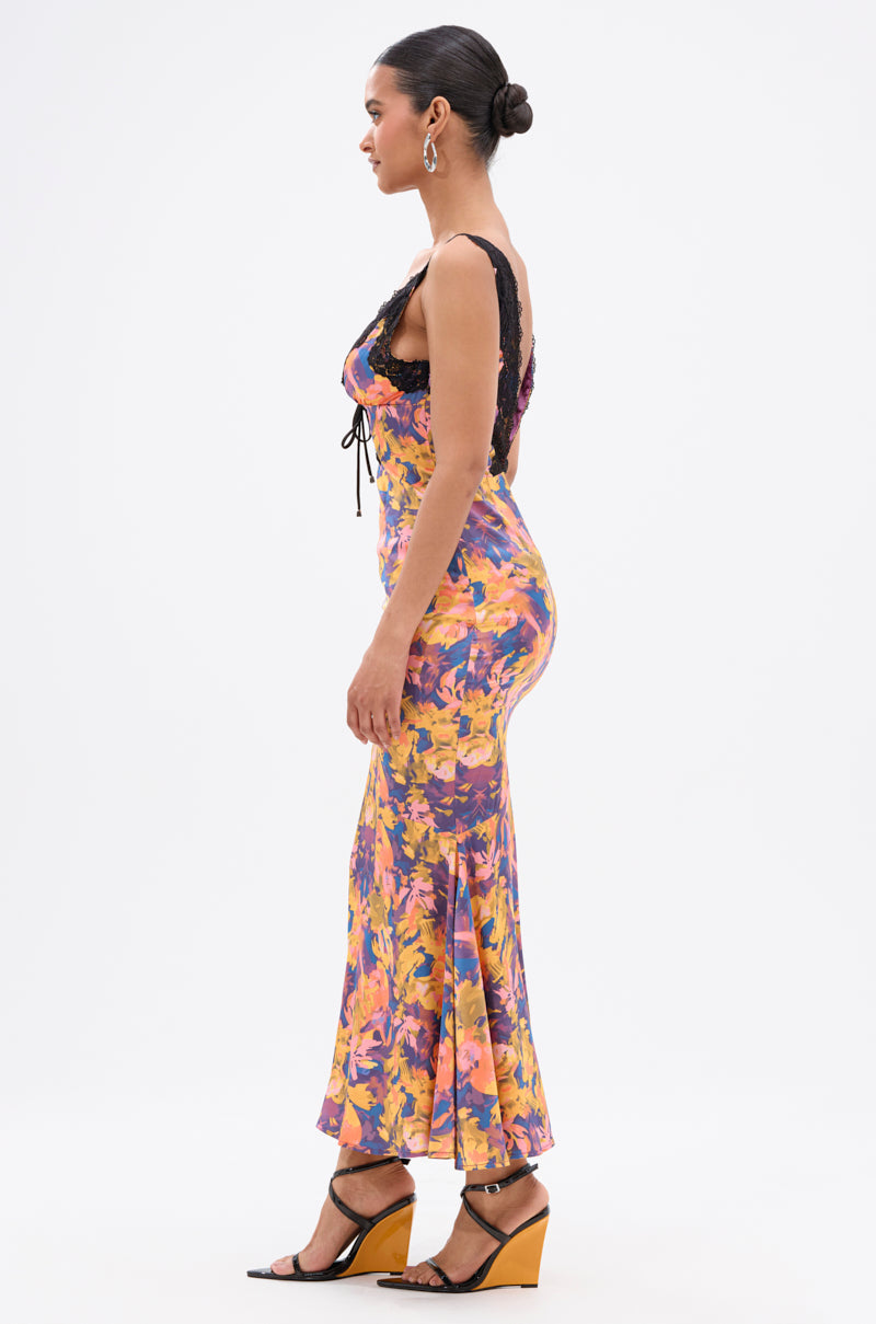 A woman in profile wears the RYAN FLORAL PRINTED SATIN MIDI DRESS, featuring a sleeveless floral design with black lace and a plunging neckline. She pairs it with wedge sandals, hoop earrings, and a neat bun against a plain white background.