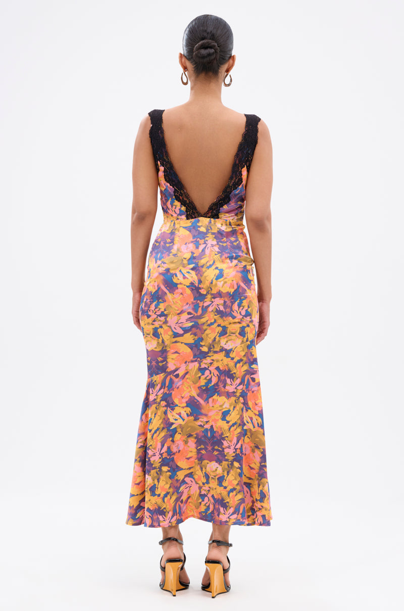 A woman faces away, wearing the RYAN FLORAL PRINTED SATIN MIDI DRESS—sleeveless, backless, with black lace trim and a vibrant orange, purple, and yellow floral print. Her hair is in a bun and she's styled with heeled sandals.