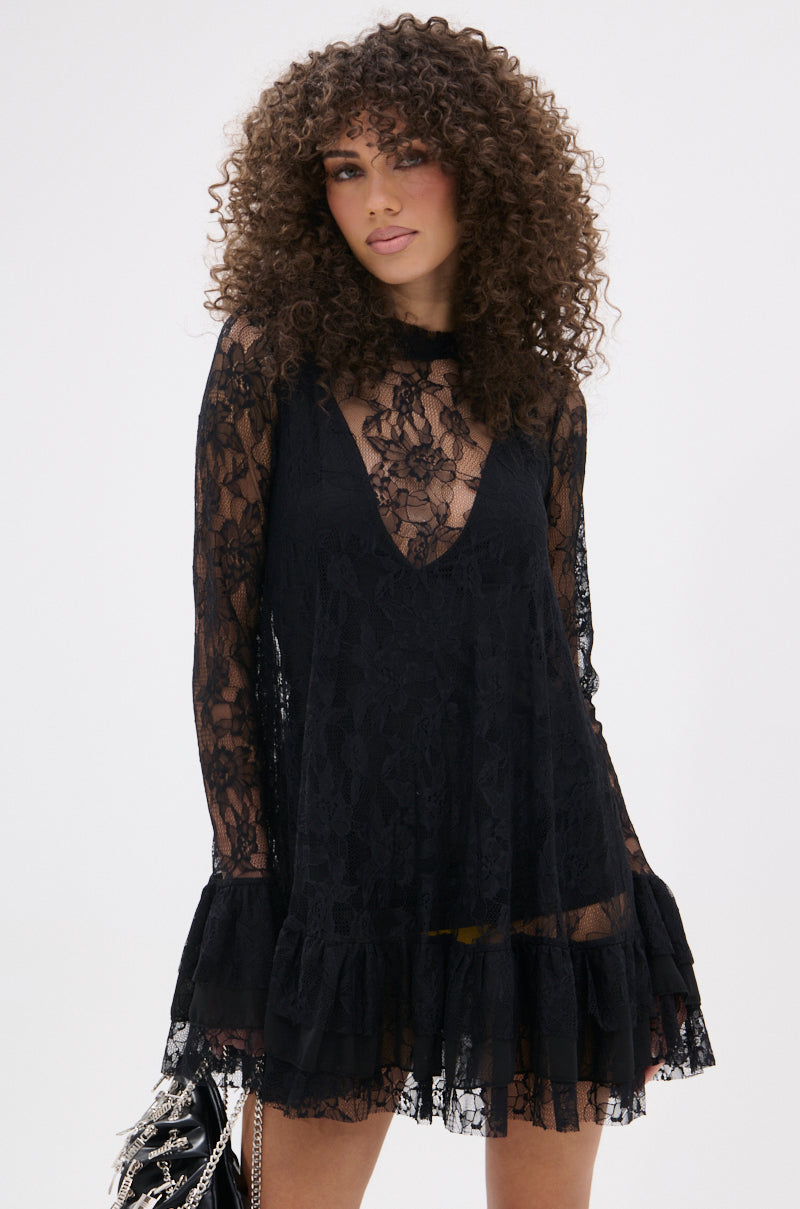 A woman with curly hair models the NEED YOU TONIGHT LACE MINI DRESS, featuring sheer floral lace, long sleeves, and a ruffled hem. She holds a black chain-detailed handbag against a plain white background.