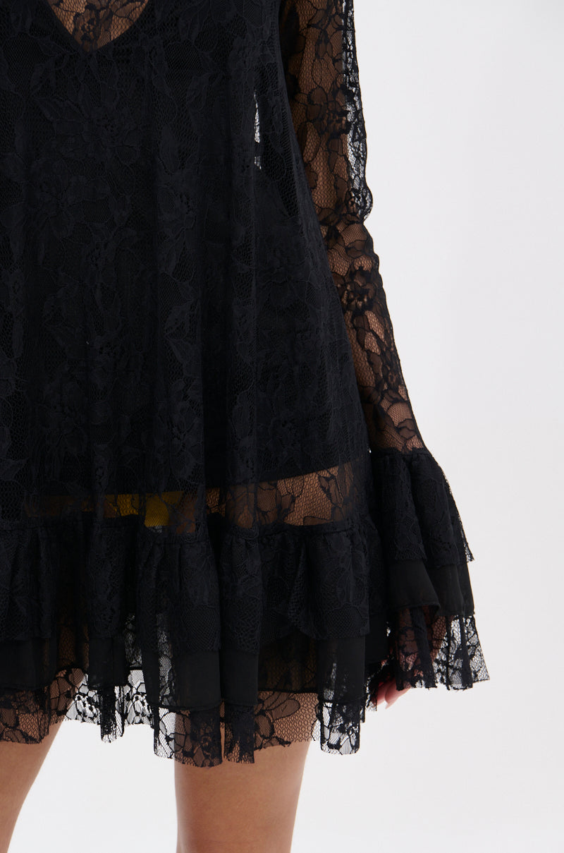 A close-up of a person wearing the NEED YOU TONIGHT LACE MINI DRESS, featuring black floral semi-sheer lace with ruffled cuffs and hem that subtly reveals their legs. The background is plain white.
