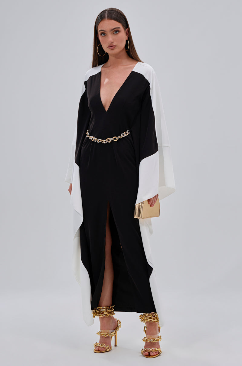 A woman with long straight hair and hoop earrings stands against a plain background, wearing the JULIE TWO TONE MIDI DRESS with a plunging V-neckline, gold chain belt, gold clutch, and gold heels.
