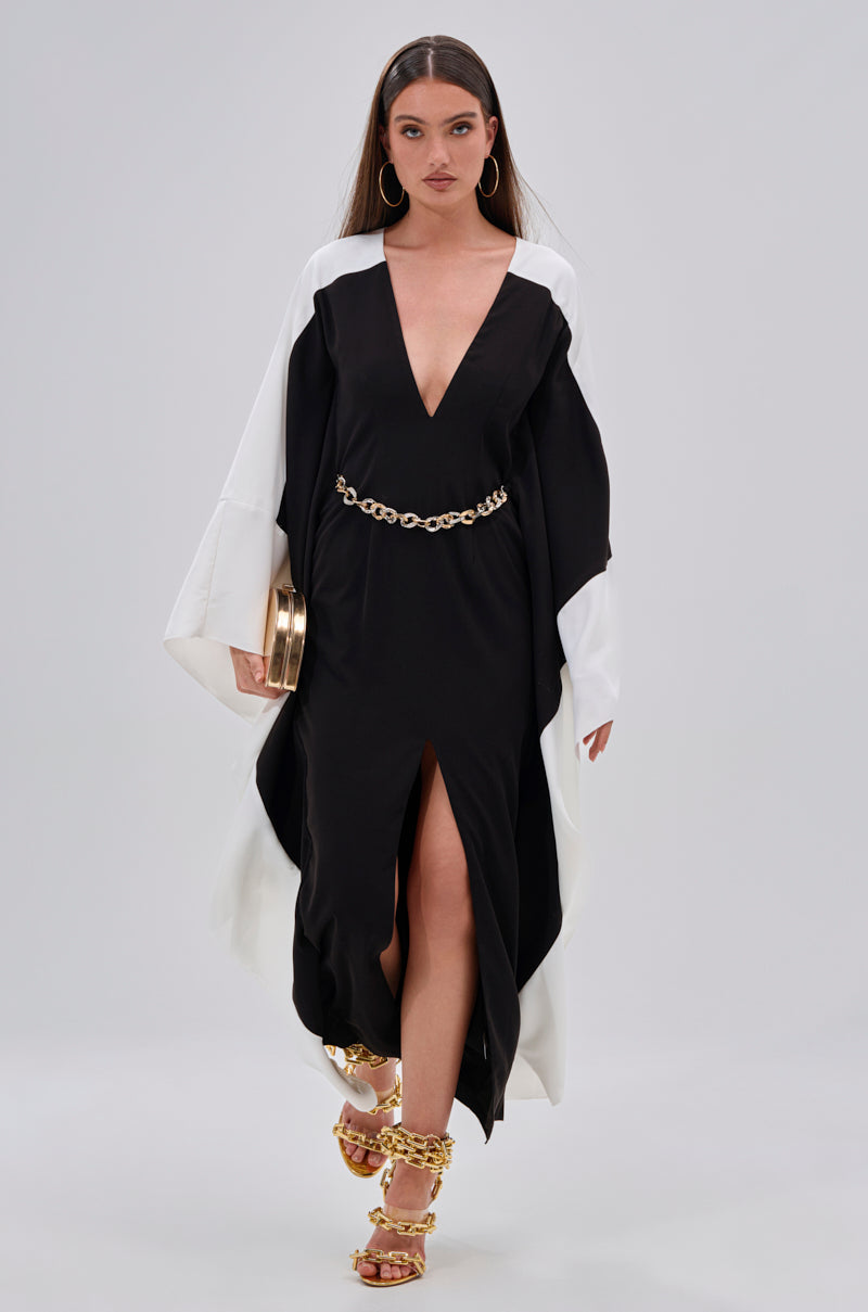 Wearing the JULIE TWO TONE MIDI DRESS with a plunging V-neckline and color block design, she styles it with gold hoops, a chain belt, chain-strap heels, and a gold clutch, gazing ahead with confidence.
