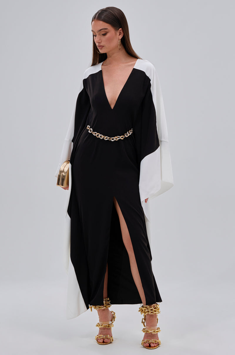 A woman in the JULIE TWO TONE MIDI DRESS with a plunging V-neckline, gold chain belt, and high slit stands against a plain background. She wears gold heeled sandals, holds a gold clutch, and looks down.