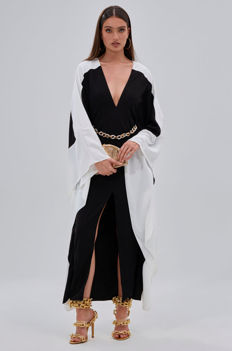 A woman stands against a plain backdrop wearing the JULIE TWO TONE MIDI DRESS with a plunging V-neckline, paired with a gold chain belt, gold high-heeled sandals, and a small clutch. Her long straight hair completes the look.