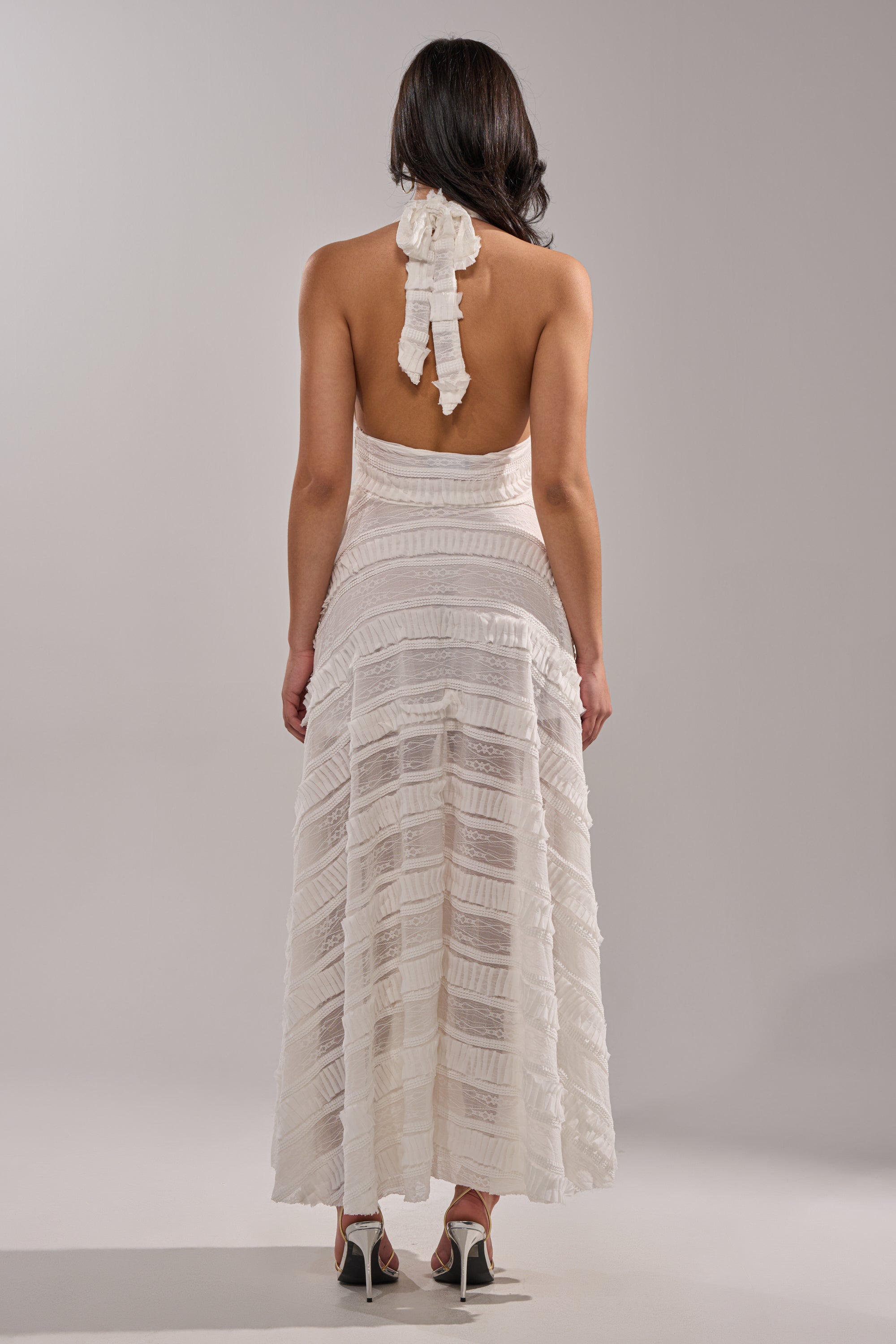 A woman with long dark hair models the SITTING PRETTY WHITE WOVEN MAXI DRESS, featuring a backless design, ruffle details, and a flowing textured skirt. She stands facing away in high heels against a plain light gray background. 4