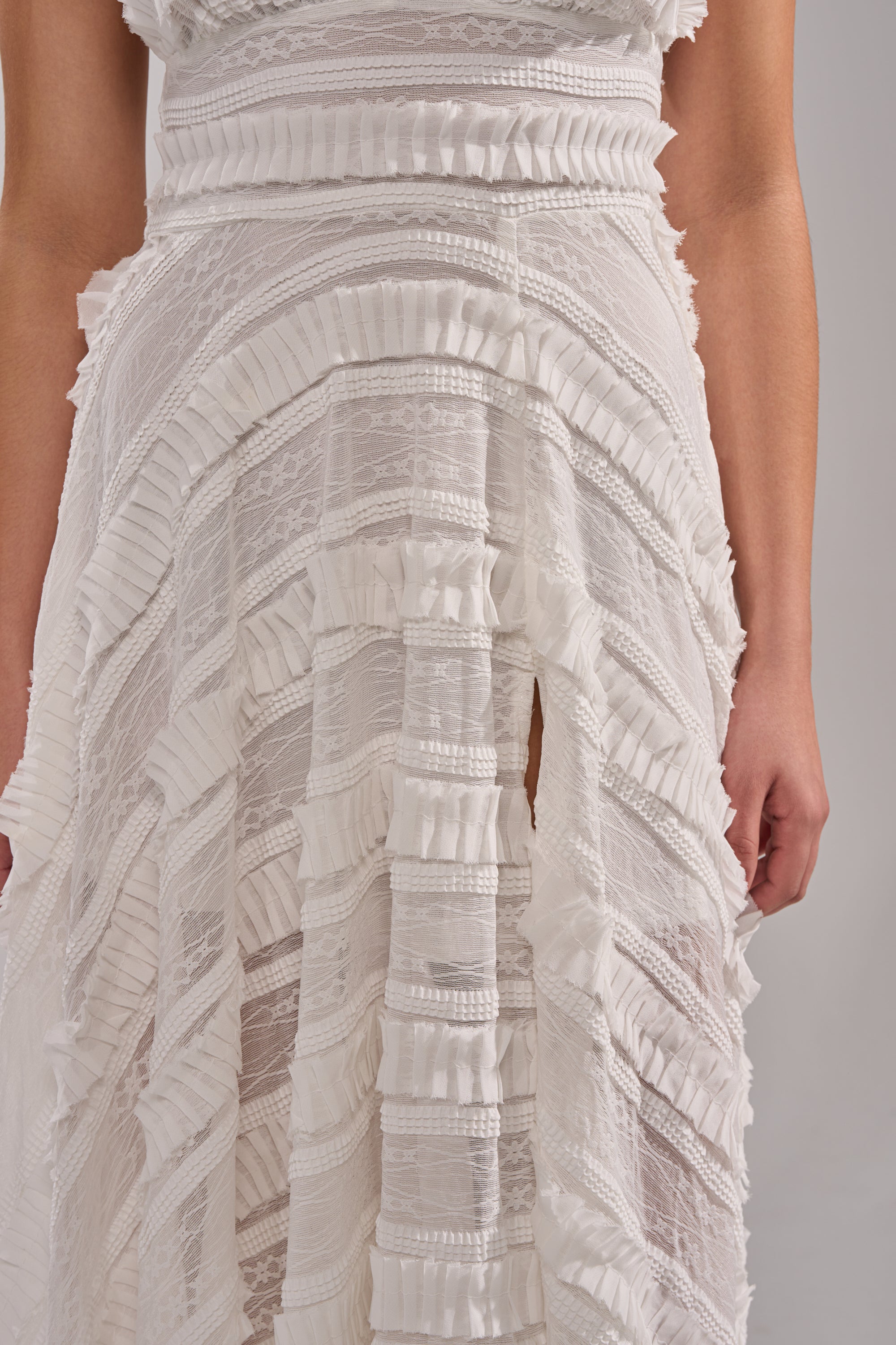 A close-up of a person wearing the SITTING PRETTY WHITE WOVEN MAXI DRESS, featuring horizontal lace panels and ruffled trim. Their arms are relaxed by their sides, highlighting the dress's intricate textures and patterns. 6