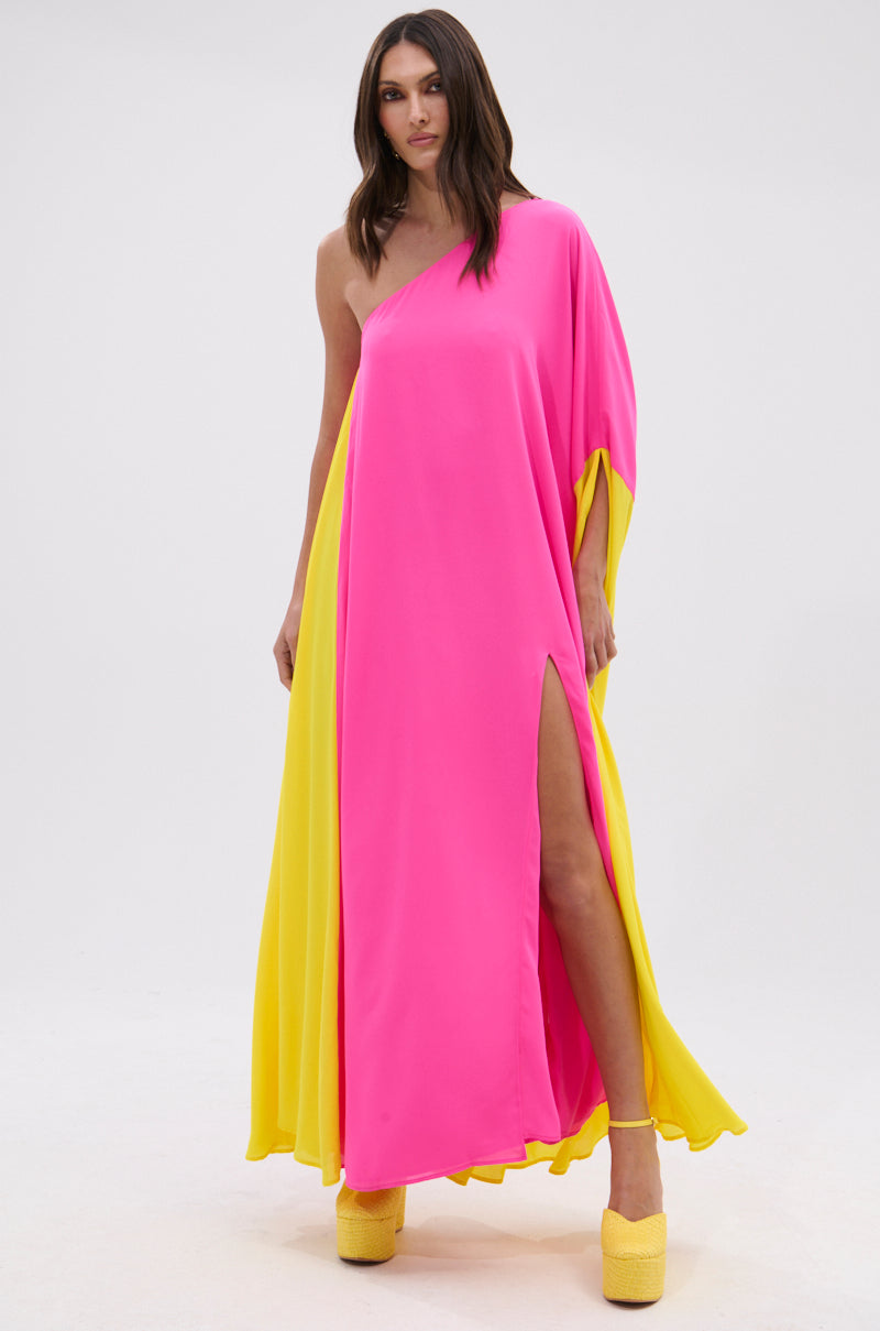 A woman stands confidently in the ONE OF A KIND MAXI DRESS, a one-shoulder maxi with fuchsia pink and yellow panels and a thigh-high slit, styled with yellow platform shoes against a plain white backdrop.