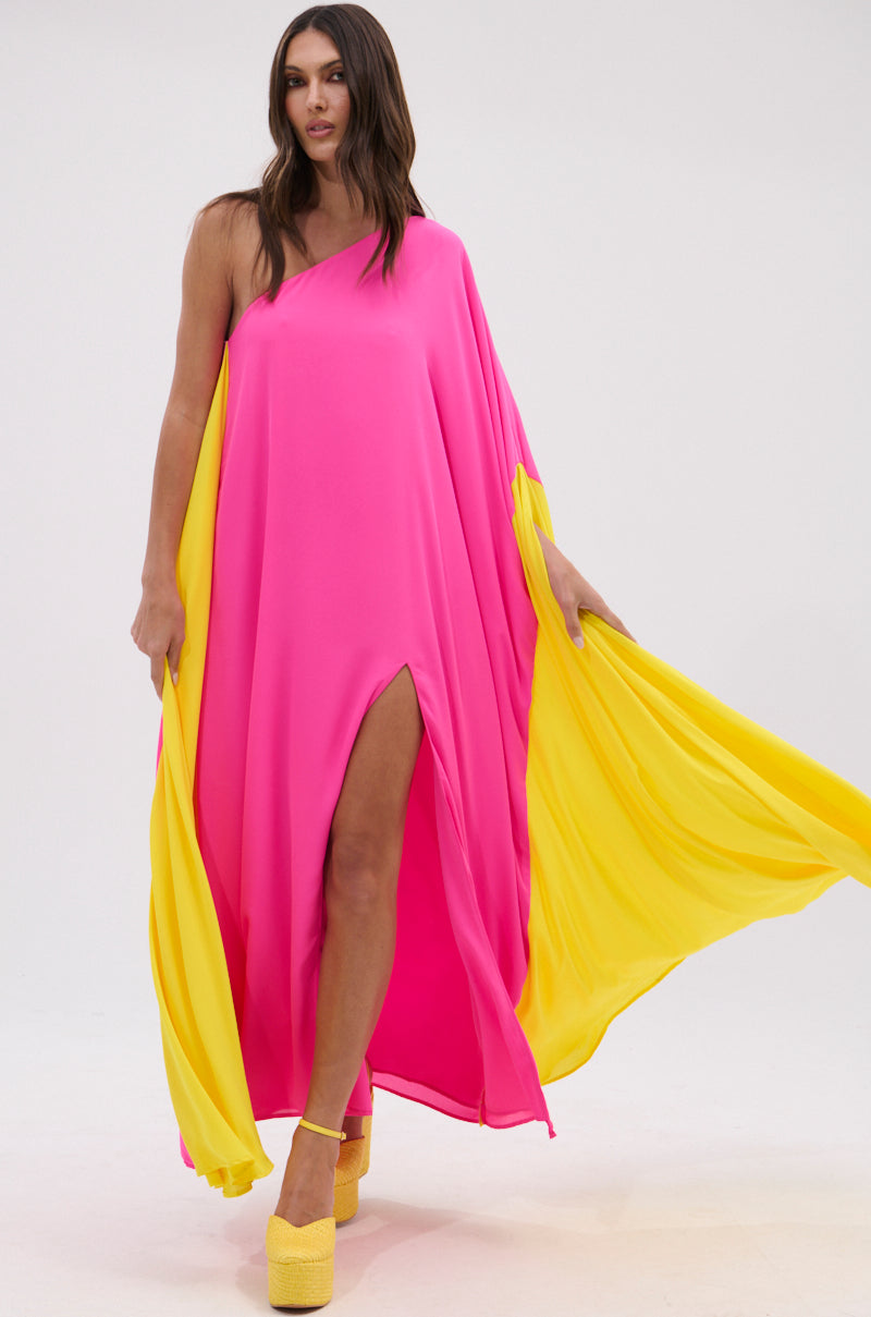 A woman poses against a plain white background wearing the ONE OF A KIND MAXI DRESS, a vibrant fuchsia pink one-shoulder maxi with flowing yellow panels, high slit, and yellow platform heels.