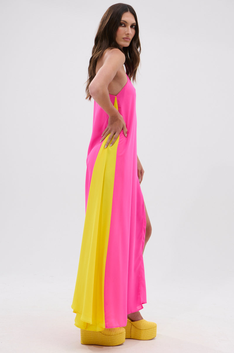 A woman with long, wavy brown hair stands sideways against a white background wearing the ONE OF A KIND MAXI DRESS in fuchsia pink and yellow, paired with platform yellow shoes, one hand on her hip.