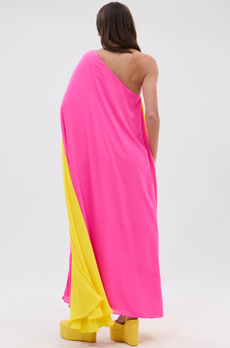 A person with long brown hair stands facing away against a plain white background, wearing the ONE OF A KIND MAXI DRESS in fuchsia pink and yellow, paired with matching yellow platform shoes.