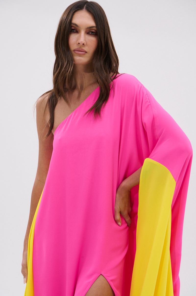 A woman with long brown hair models the ONE OF A KIND MAXI DRESS, a fuchsia pink and yellow one-shoulder maxi with a loose sleeve and high slit, standing against a plain light background.