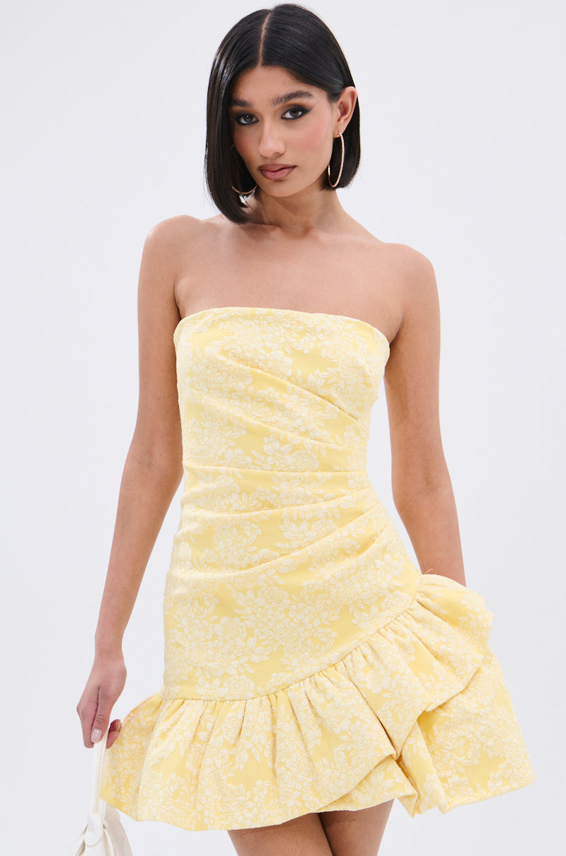 A woman with straight dark hair models the GARDEN PARTY MINI DRESS, showcasing its floral print and ruched bodice. She pairs it with large hoop earrings and a white handbag.