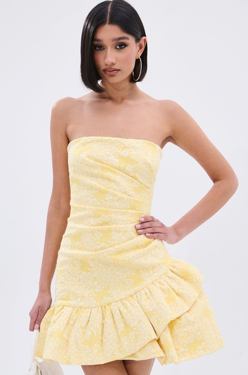 A woman poses with one hand on her hip against a white background, wearing the GARDEN PARTY MINI DRESS—a strapless yellow mini dress with a white floral print, ruched bodice, and ruffled hem.