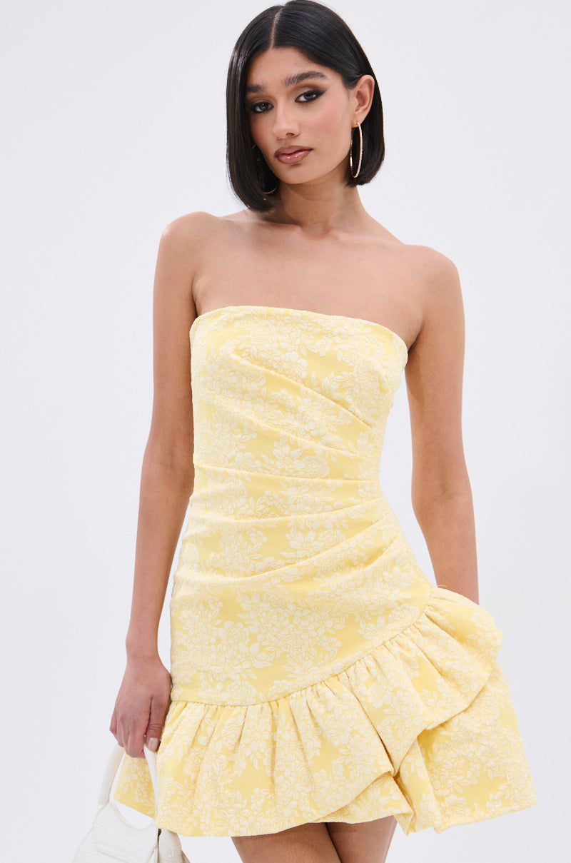 A woman with straight, shoulder-length dark hair wears the GARDEN PARTY MINI DRESS, a strapless yellow mini dress with a white floral print and ruffled hem, styled with gold hoop earrings and a white handbag against a plain white background.