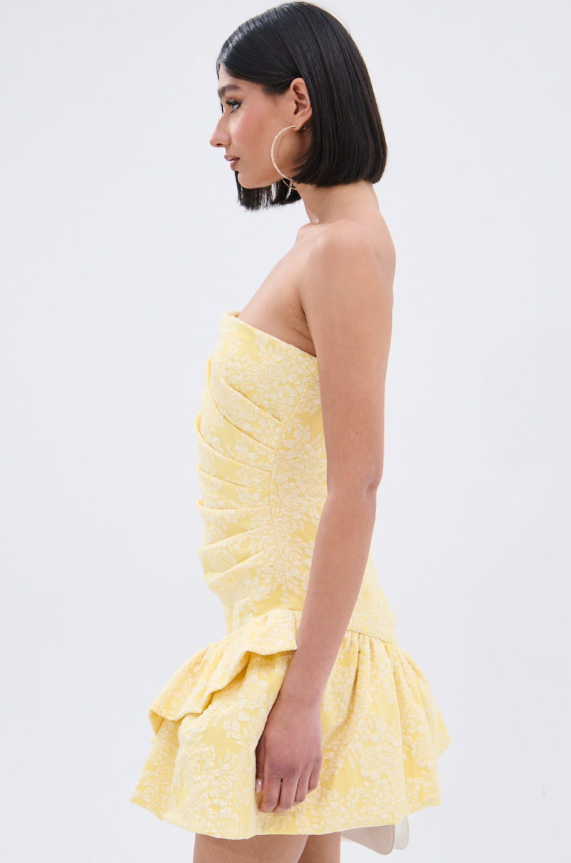 A woman with straight, shoulder-length dark hair and hoop earrings wears the strapless GARDEN PARTY MINI DRESS, which features a ruched bodice and ruffled hem. She is standing in profile against a plain light background.
