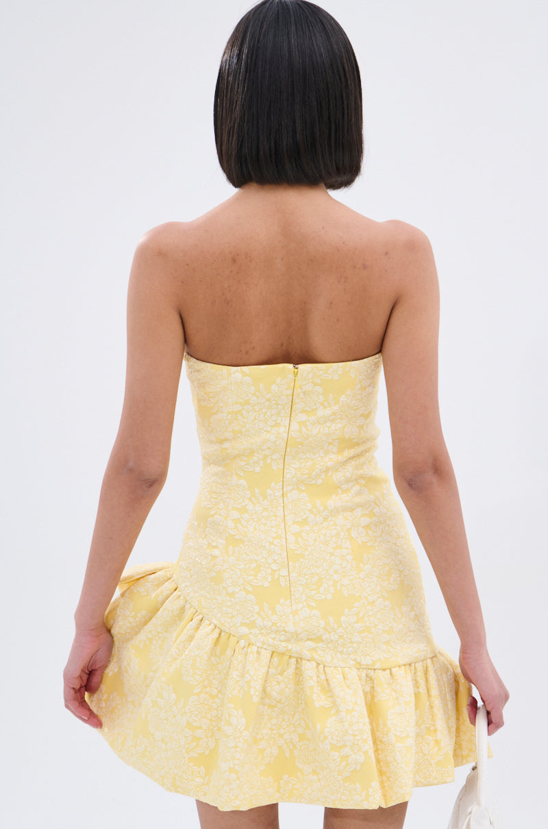 A person with short dark hair is seen from behind in the GARDEN PARTY MINI DRESS, which has a strapless ruched bodice, yellow floral print, and ruffled hem. They hold the sides of the dress against a plain, light background.