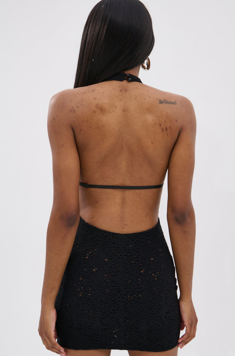 A woman with straight long hair and a script tattoo on her upper right shoulder stands with her back to the camera, wearing the ON THE MOVE BODYCON MINI DRESS IN BLACK featuring a halter neckline and openwork crochet lace. The background is plain and light.