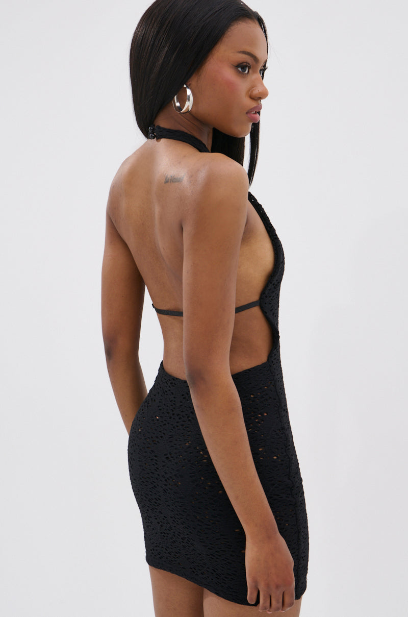 A woman models the ON THE MOVE BODYCON MINI DRESS IN BLACK, a sleeveless, backless mini dress, paired with large silver hoop earrings, as she stands sideways and looks over her shoulder against a plain light background.