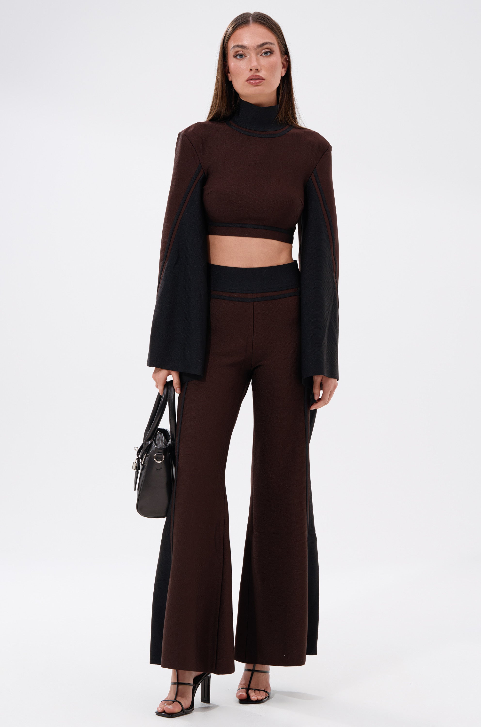 A woman stands against a white background wearing the MAZIE WIDE LEG PALAZZO PANT with a matching dark brown, long-sleeve crop top featuring flared sleeves. She accessorizes with a black handbag and black heeled sandals.