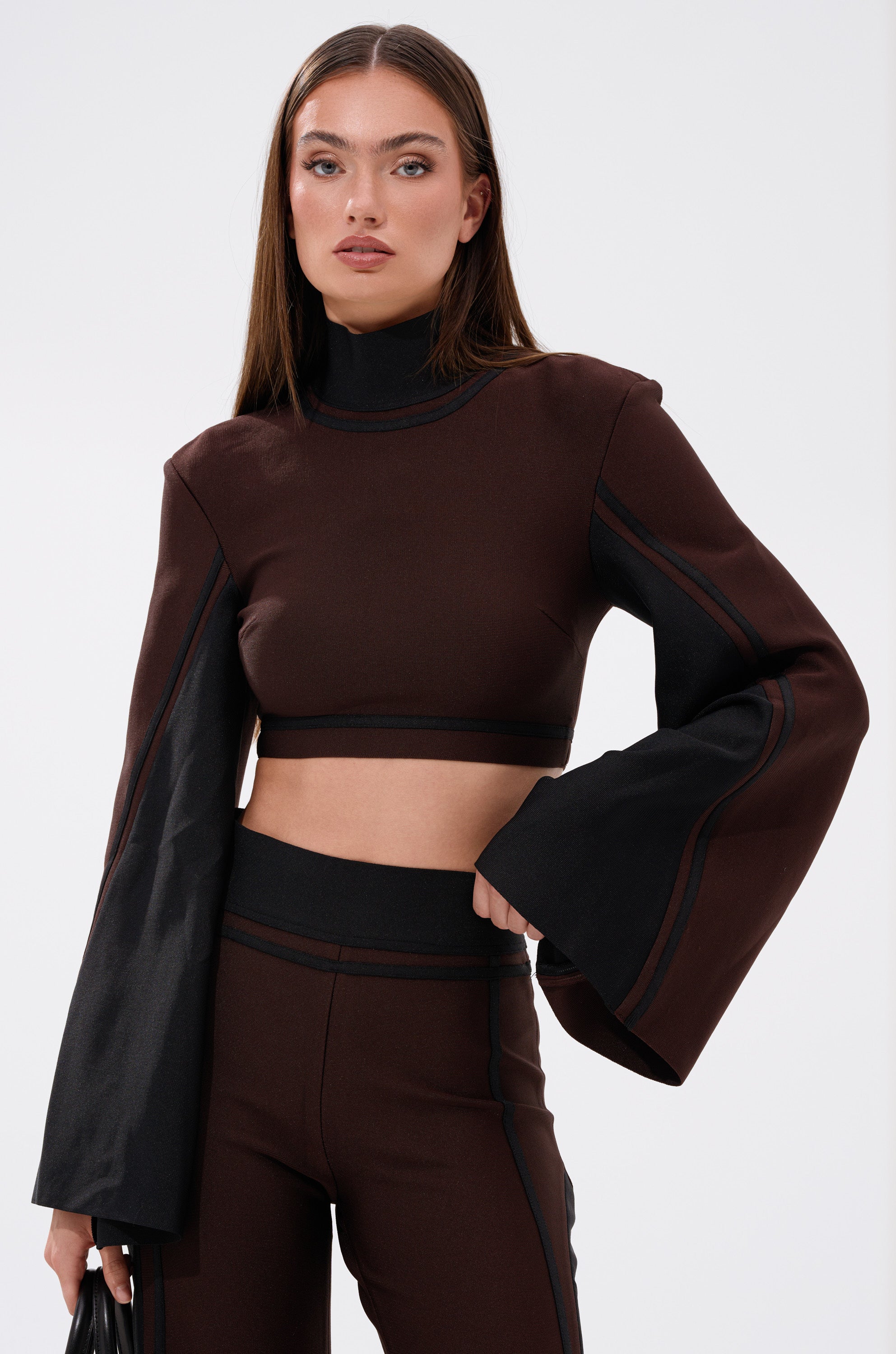 A woman poses confidently against a plain, light background wearing the MAZIE MOCK NECK SWEATER with flared sleeves, paired with matching high-waisted pants.