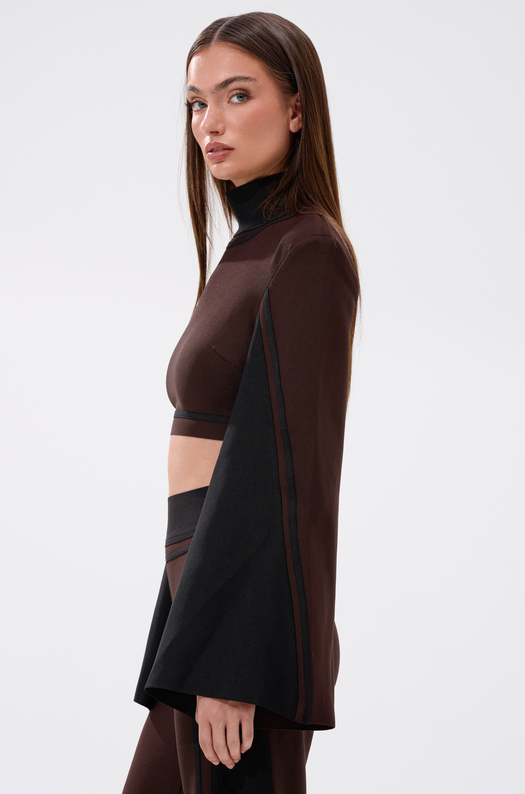 A woman with long brown hair models the MAZIE MOCK NECK SWEATER, a brown cropped style with wide sleeves and matching high-waisted pants, standing in profile against a plain white background.