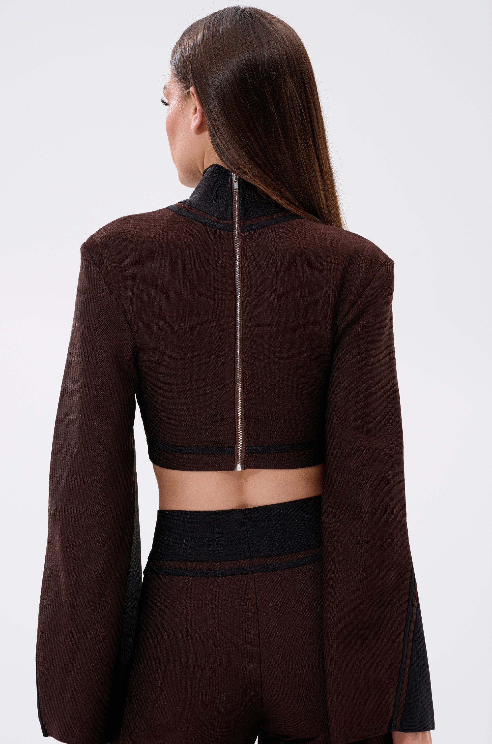 A woman with straight brown hair is seen from behind against a light background, wearing the MAZIE MOCK NECK SWEATER—a dark brown, cropped design with bell sleeves and a visible back zipper.