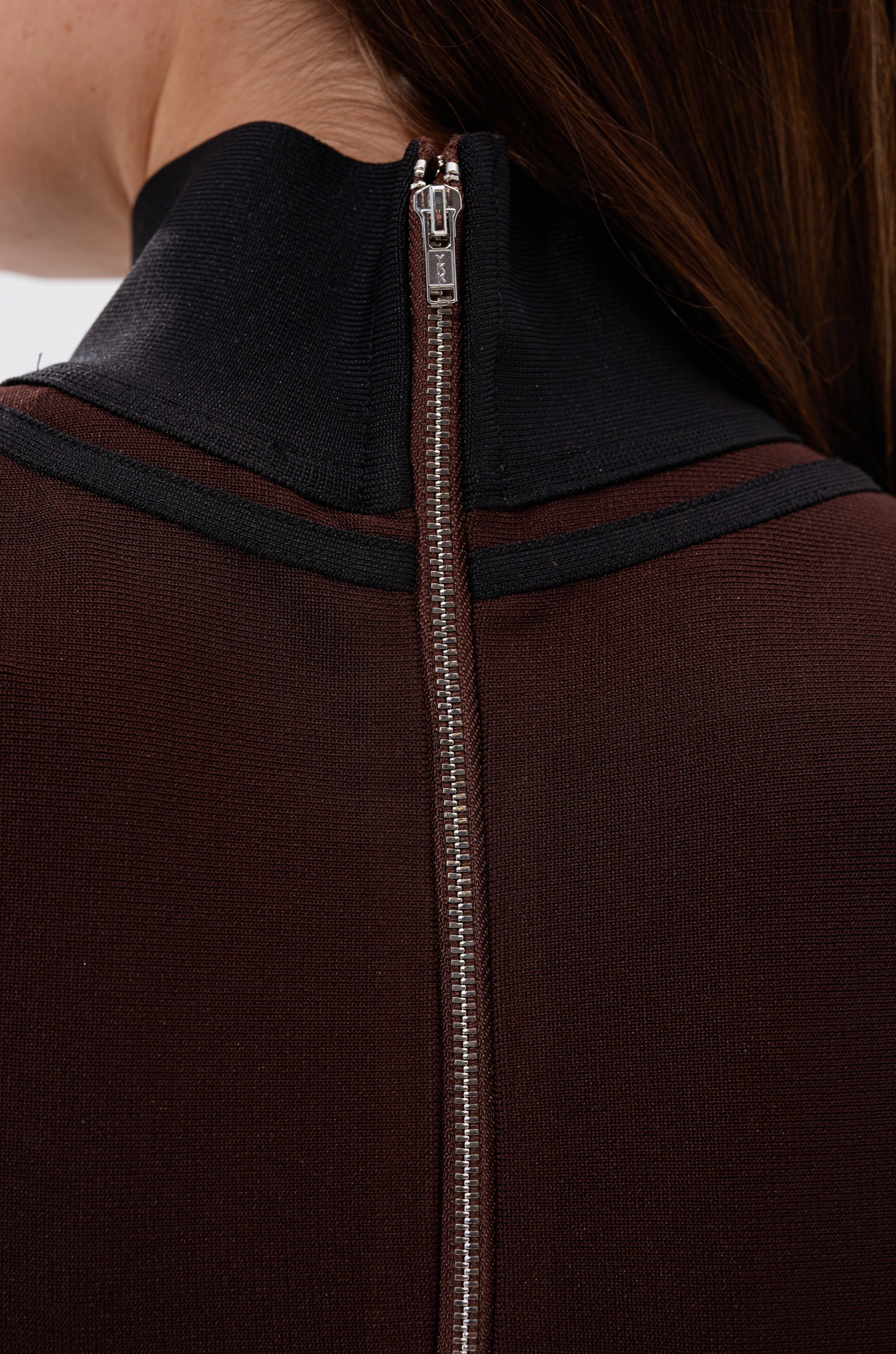 Close-up of the back of a person’s neck and upper back, wearing the MAZIE MOCK NECK SWEATER in brown and black with a silver center zipper. The person’s brown hair is partially visible.