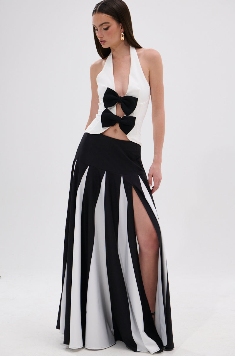 A woman wears the RACHEL SATIN MAXI SKIRT, a monochrome high-slit piece paired with a sleeveless black and white gown featuring a plunging neckline and two large bows on the bodice. 0