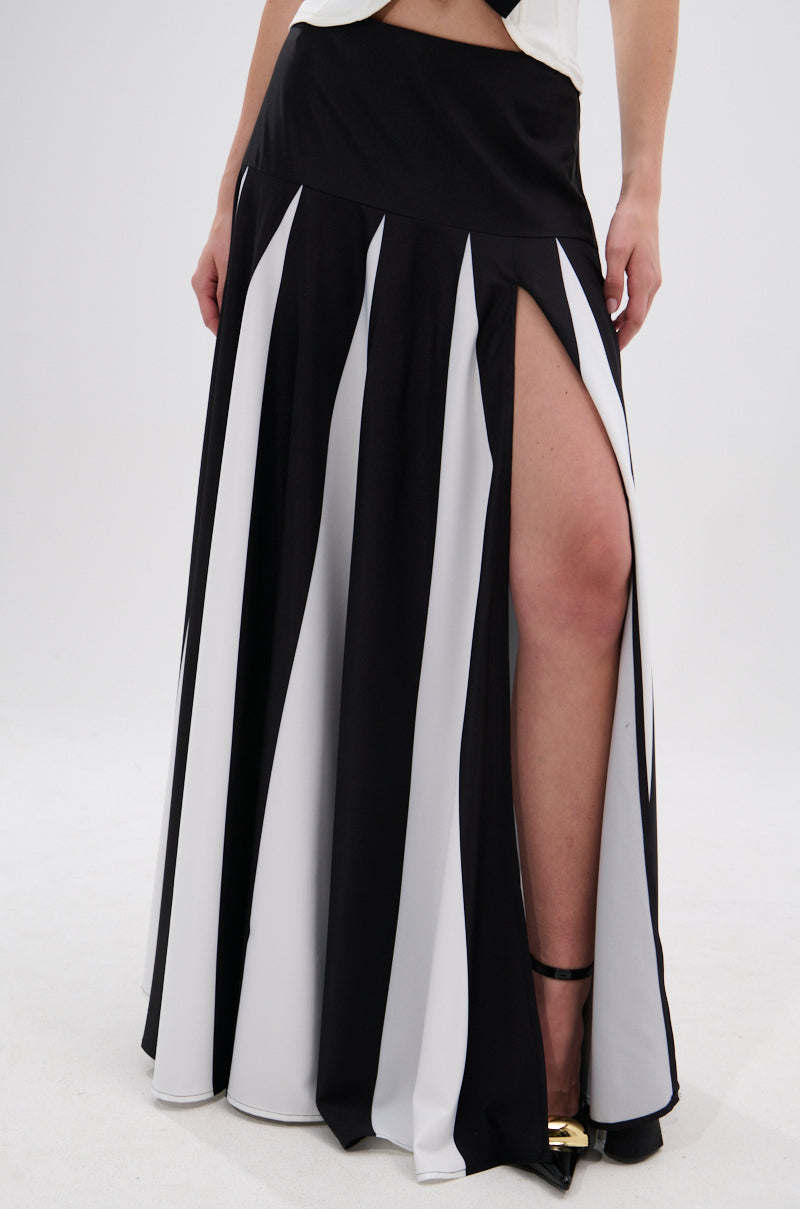 A person wears the RACHEL SATIN MAXI SKIRT, a black and white striped satin maxi with a high-rise waistband and side slit. Black ankle-strap high heels complete the look, with only part of the upper body visible. 1