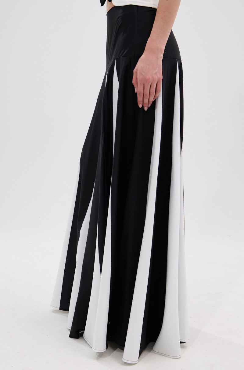 A person wears the RACHEL SATIN MAXI SKIRT, featuring bold black and white vertical stripes, a high-rise waistband, and a flowing floor-length silhouette. Their hand rests by their side against a plain white background. 3
