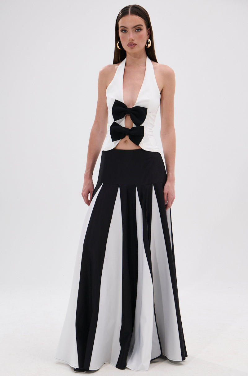 A woman standing against a plain white background wears the Rachel Satin Maxi Skirt, featuring a long monochrome design with a high-rise waistband, paired with a sleeveless halter top and large black bow at the bodice. 6