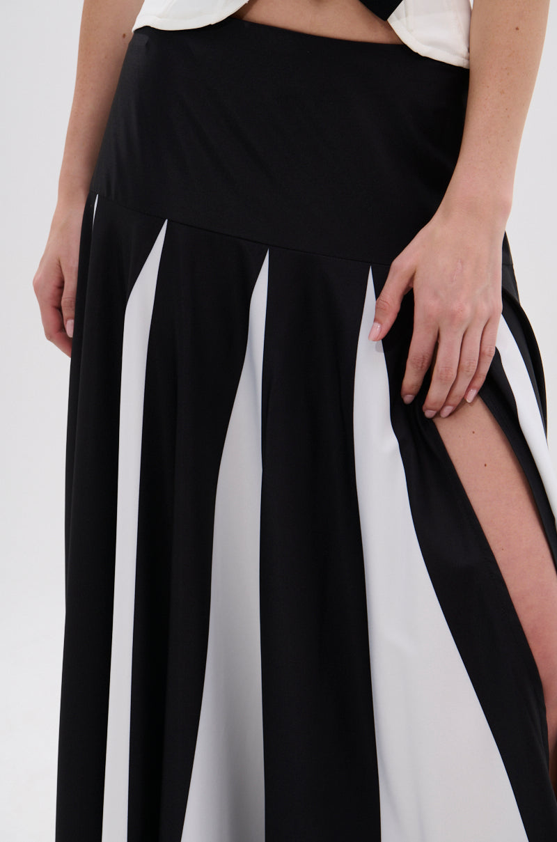 A person wears the RACHEL SATIN MAXI SKIRT featuring black and white vertical stripes, a high slit revealing their leg, and a high-rise waistband. Their hands rest on the waistband. The background is plain and light-colored. 8