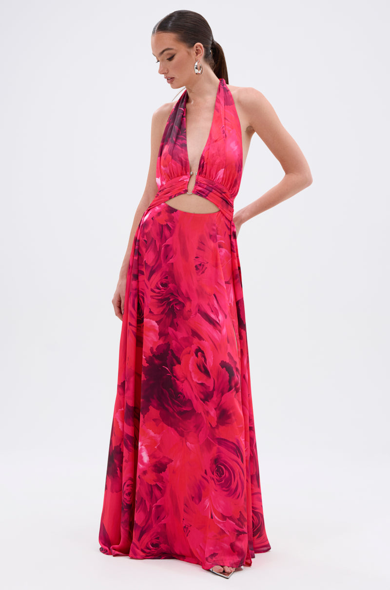 A woman models the ALL EYES ON ME CUT OUT MAXI DRESS, featuring a vibrant red and pink floral print, halter neckline, front cutout, deep neckline, and flowing skirt against a plain white background.