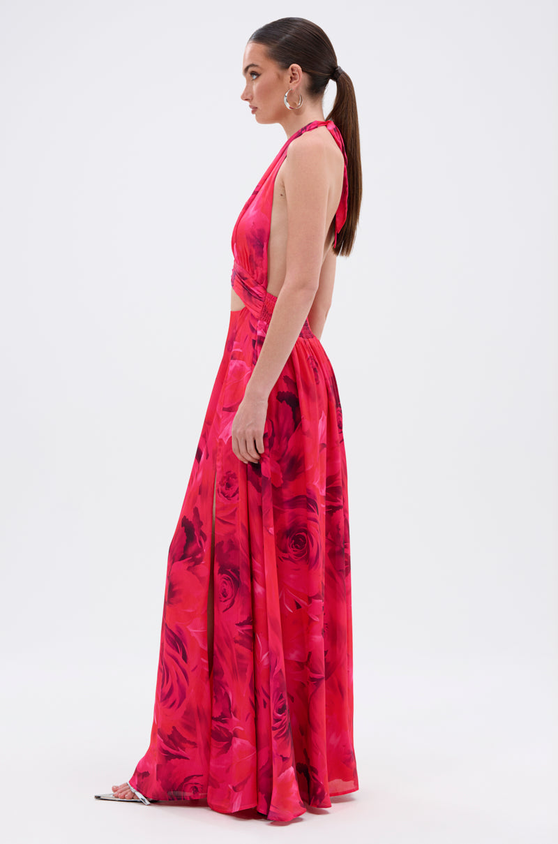 A woman stands in profile wearing the ALL EYES ON ME CUT OUT MAXI DRESS, a sleeveless, backless floral halter maxi with side cutouts featuring red and pink flowers. She styles it with a low ponytail, hoop earrings, and open-toed shoes.