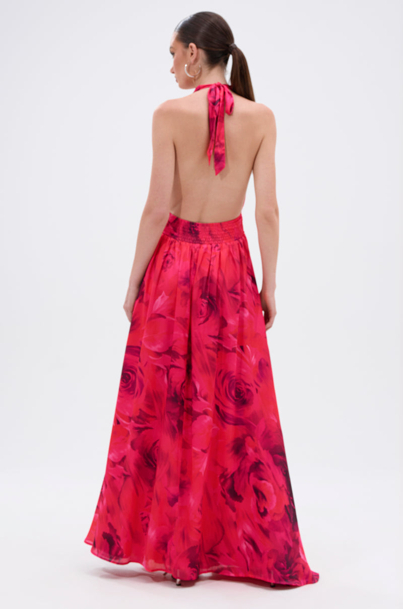 A woman with a sleek ponytail is seen from behind in the ALL EYES ON ME CUT OUT MAXI DRESS, featuring a vibrant pink and red floral print, halter neckline, and a flowing, floor-length skirt that enhances her elegant look.