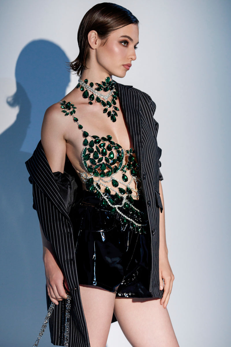 A woman poses sideways against a softly lit background, wearing the LUCKY ME RHINESTONE BLOUSE with emerald green rhinestones, a pinstripe blazer draped over her shoulders, and black shiny shorts. Her hair is slicked back. 0