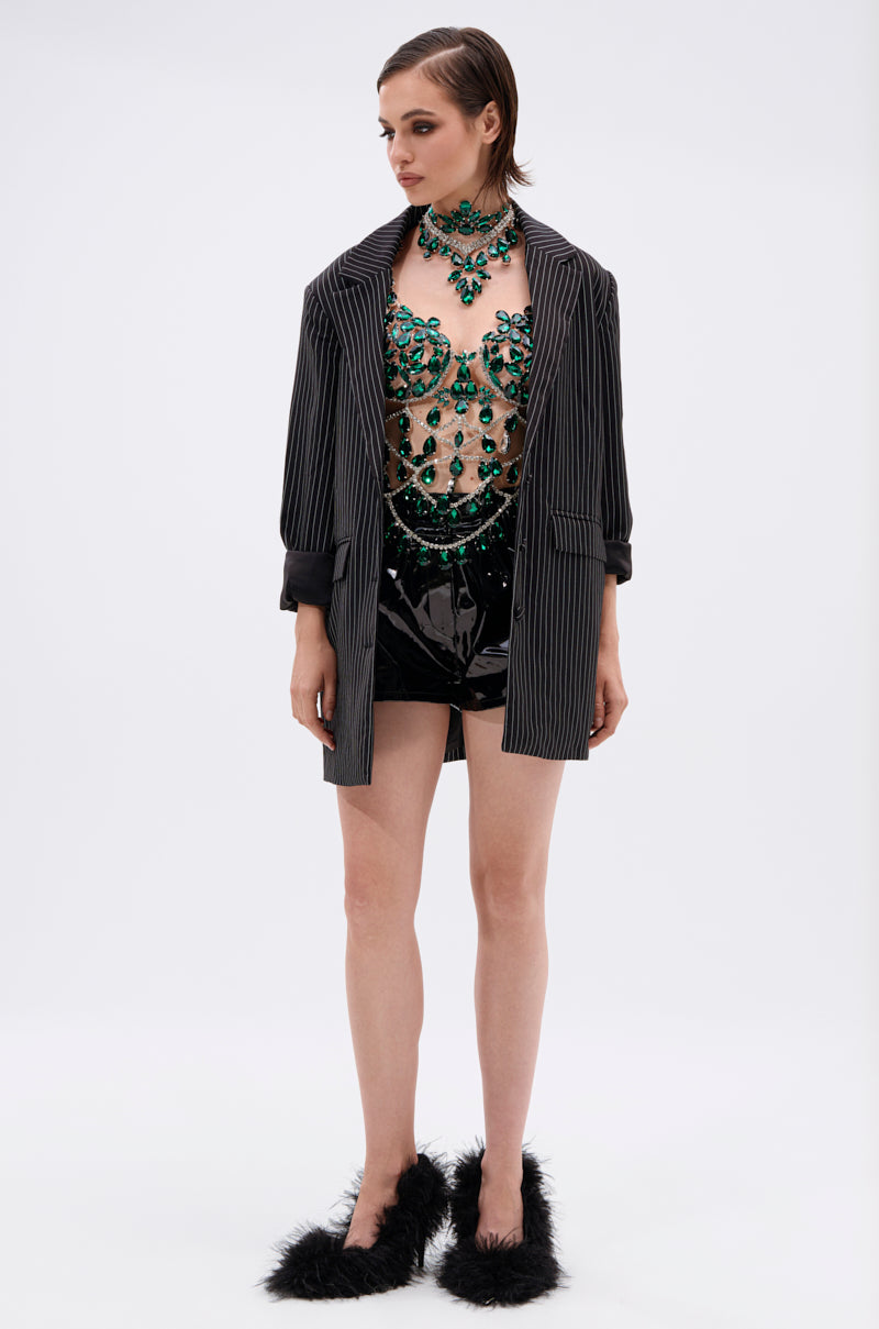 A woman stands against a plain background wearing the LUCKY ME RHINESTONE BLOUSE, a pinstripe blazer, shiny black shorts, and black fuzzy high-heeled shoes. 7