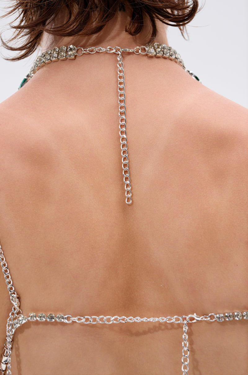 Close-up of a person with short brown hair wearing the LUCKY ME RHINESTONE BLOUSE featuring layered silver chains and emerald green rhinestone accents draped around the upper back and shoulders, set against a plain light background. 8