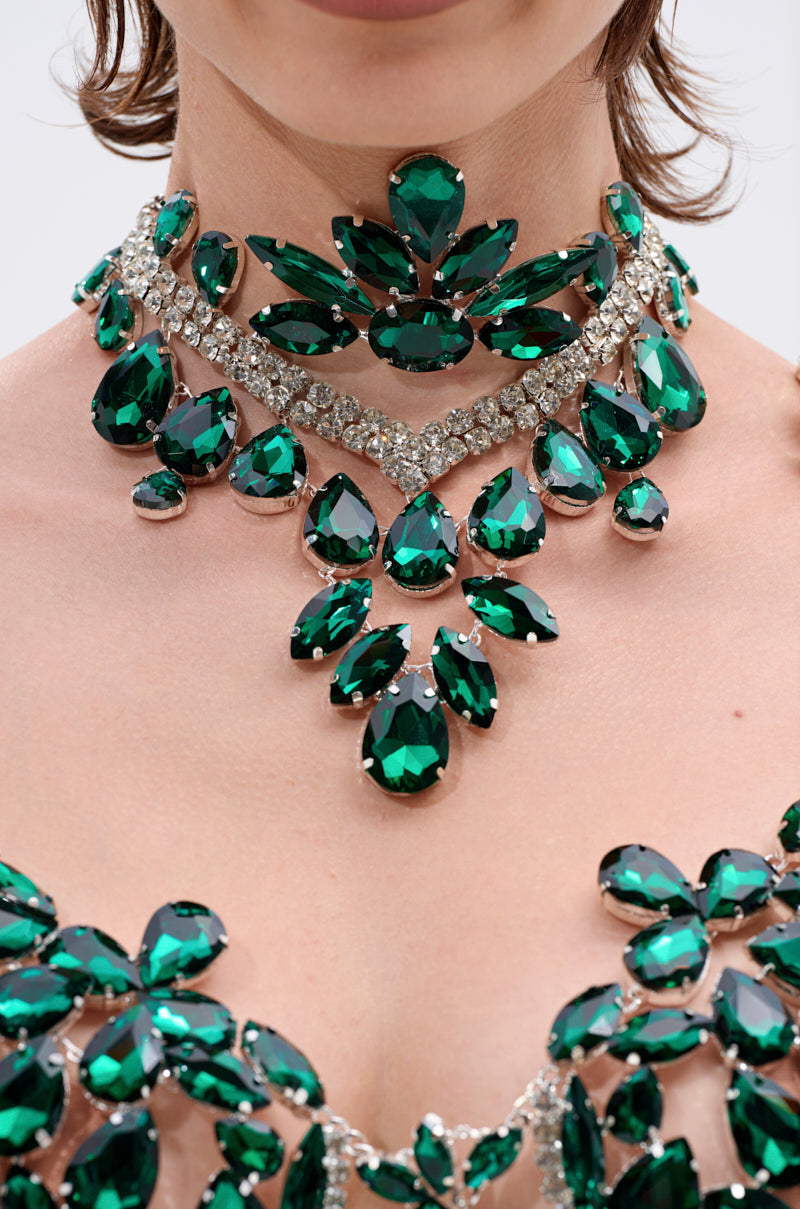 A close-up of a person wearing the LUCKY ME RHINESTONE BLOUSE, featuring ornate emerald green rhinestones and clear crystals in intricate patterns around the neck and chest. 9