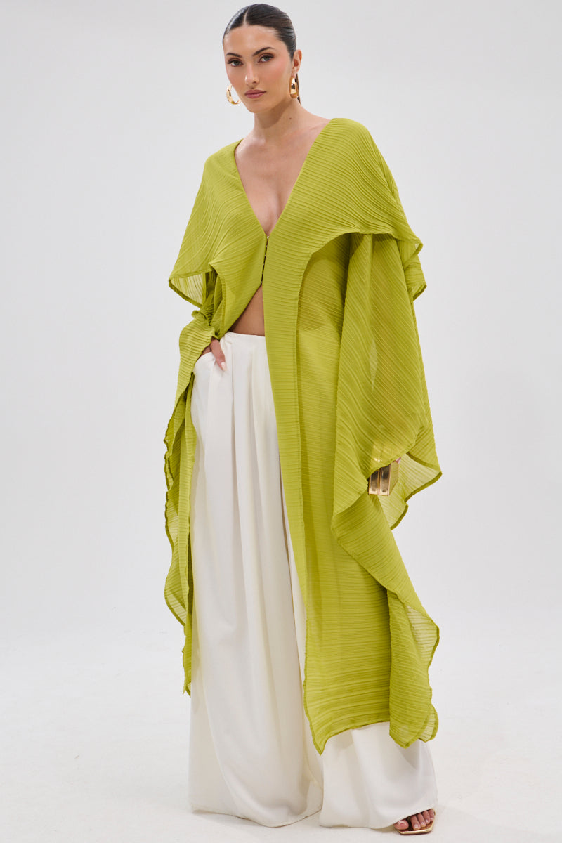 A woman models the FLY AWAY WITH ME BLOUSE IN CHARTREUSE, a pleated longline poncho with a deep V neckline, paired with white wide-leg pants and gold drop earrings. Her hair is pulled back against a plain white background. 1