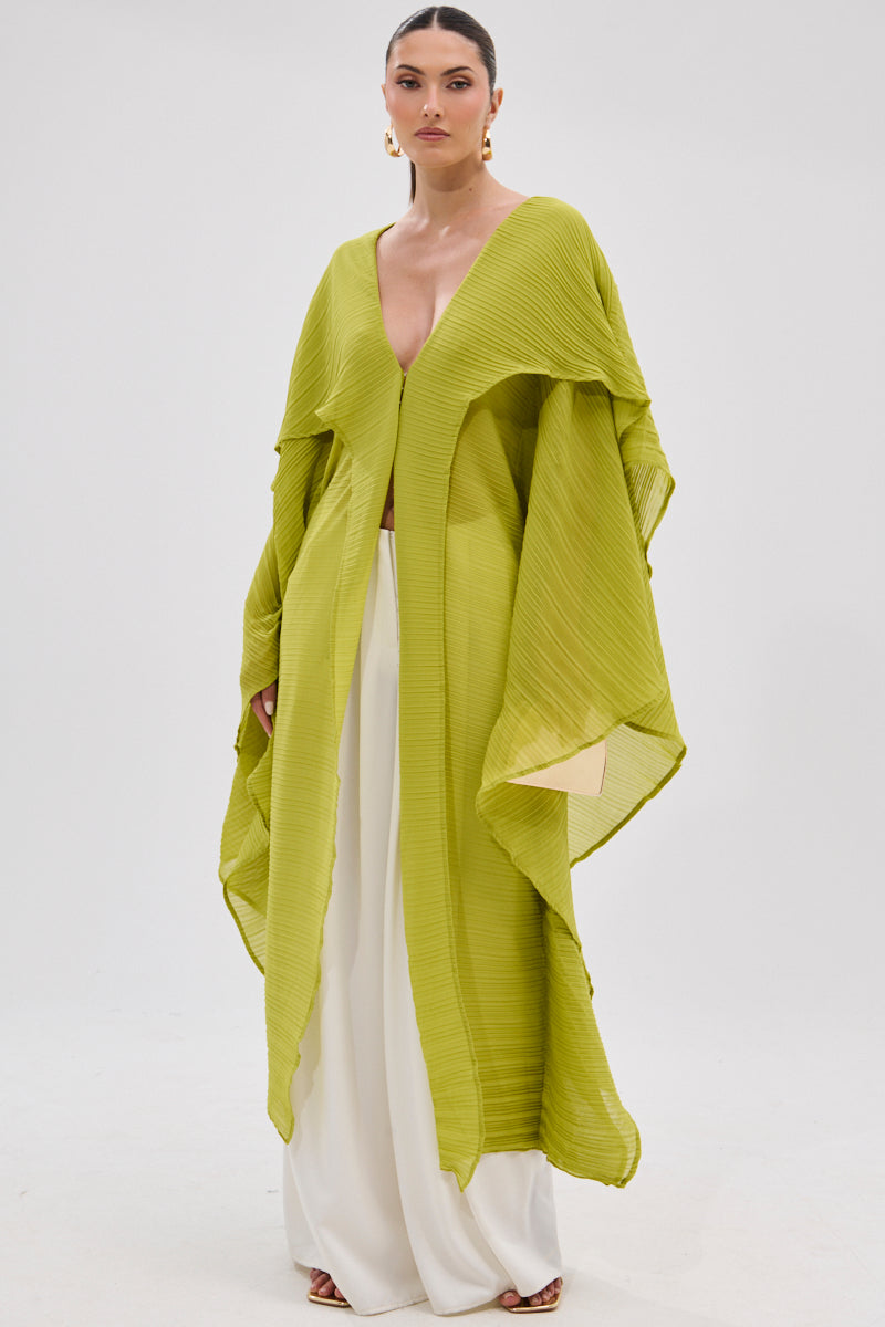 A woman stands against a white background wearing the FLY AWAY WITH ME BLOUSE IN CHARTREUSE, featuring dramatic sleeves and a longline poncho silhouette, paired with wide-leg white pants and gold hoop earrings. Her hair is sleek and pulled back. 2