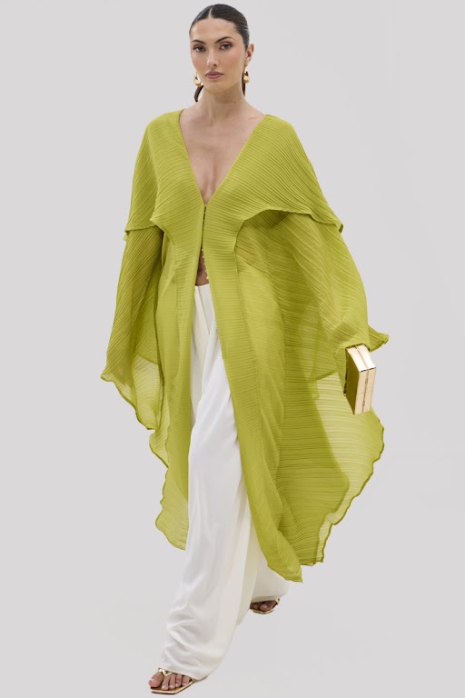 A woman in the FLY AWAY WITH ME BLOUSE IN CHARTREUSE styled as a flowing, longline cape over white wide-leg pants walks forward with a gold book clutch, hair pulled back and a confident look against a plain light background. 3